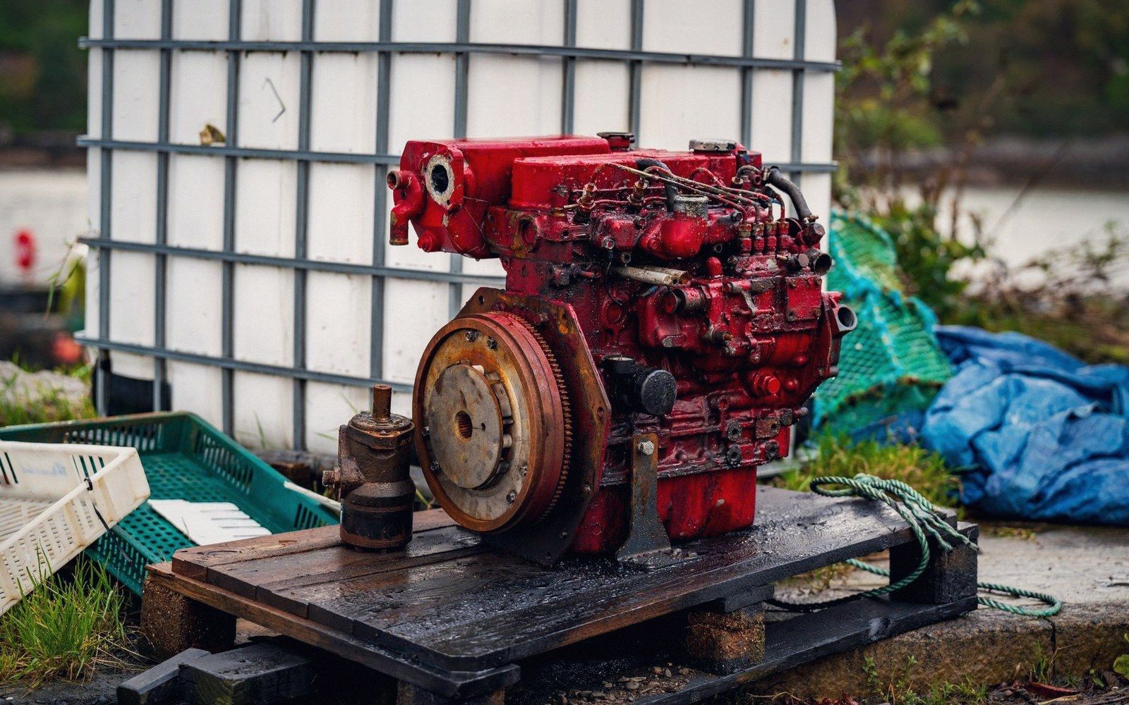 A red marine diesel engine with exposed flywheel sits on a wooden pallet outdoors